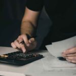 Manos usando una calculadora y revisando documentos financieros para calcular costos y gastos del negocio.
