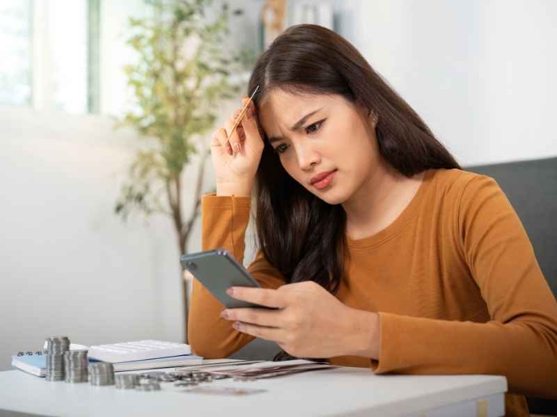Mujer preocupada mirando el celular junto a monedas y papeles, reflejando estrés por falta de control financiero.