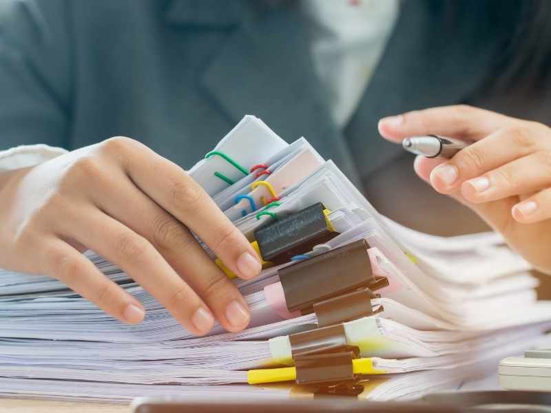 Primer plano de manos organizando una gran montaña de documentos con clips de colores, simbolizando la carga administrativa y el exceso de trámites.