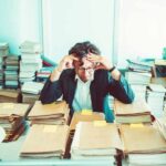 Hombre de negocios abrumado y estresado rodeado de pilas de carpetas y documentos, ilustrando el impacto emocional del costo de la burocracia.