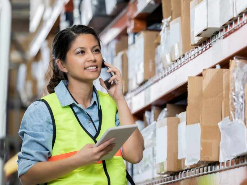 Trabajadora en almacén revisando inventario con tablet entre estanterías y cajas.