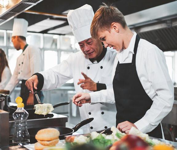 Cocinero experimentado enseñando técnicas de preparación de alimentos a un chef joven en la cocina de un restaurante.