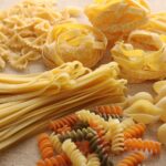 Diferentes tipos de pasta seca, como espaguetis, fusilli, penne y otras variedades, organizados sobre una superficie de madera.