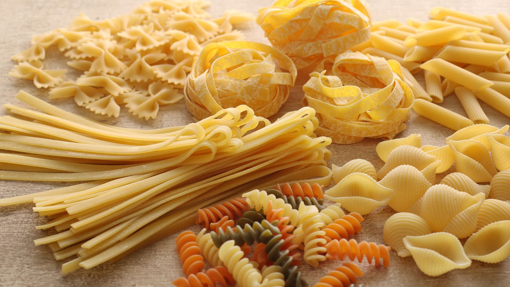 Diferentes tipos de pasta seca, como espaguetis, fusilli, penne y otras variedades, organizados sobre una superficie de madera.