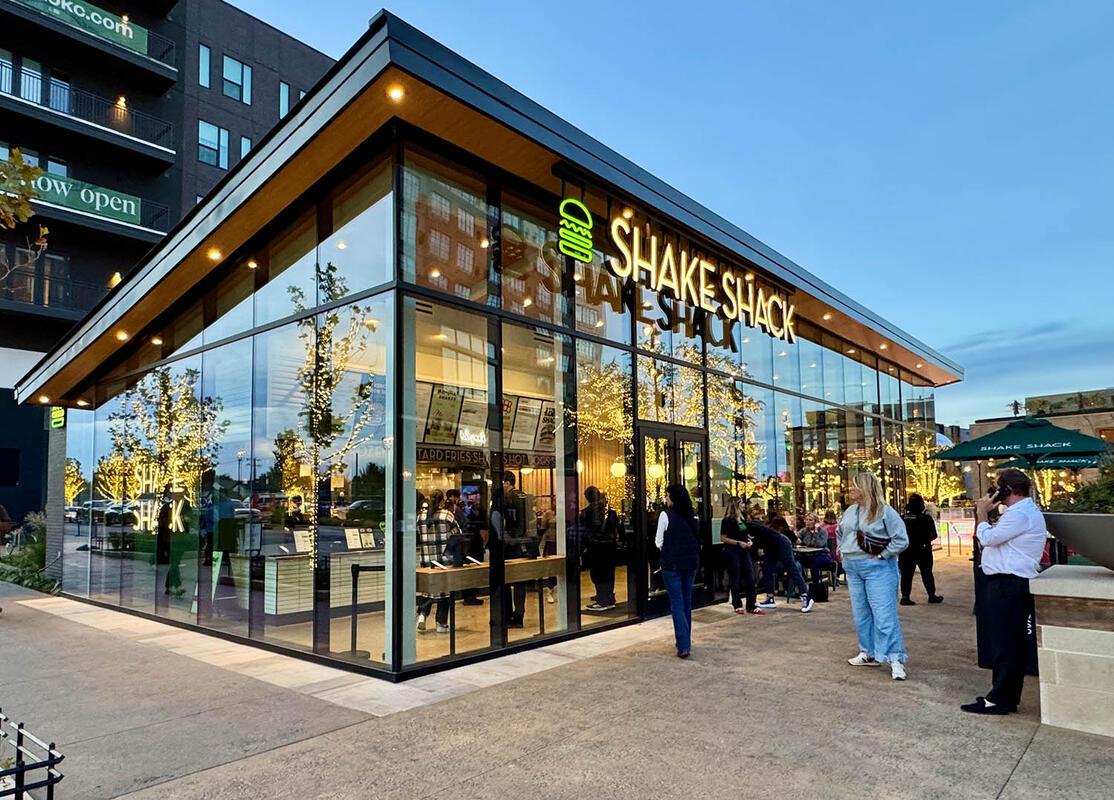 Fachada de un restaurante moderno con grandes ventanales, mesas al aire libre y clientes entrando al establecimiento.
